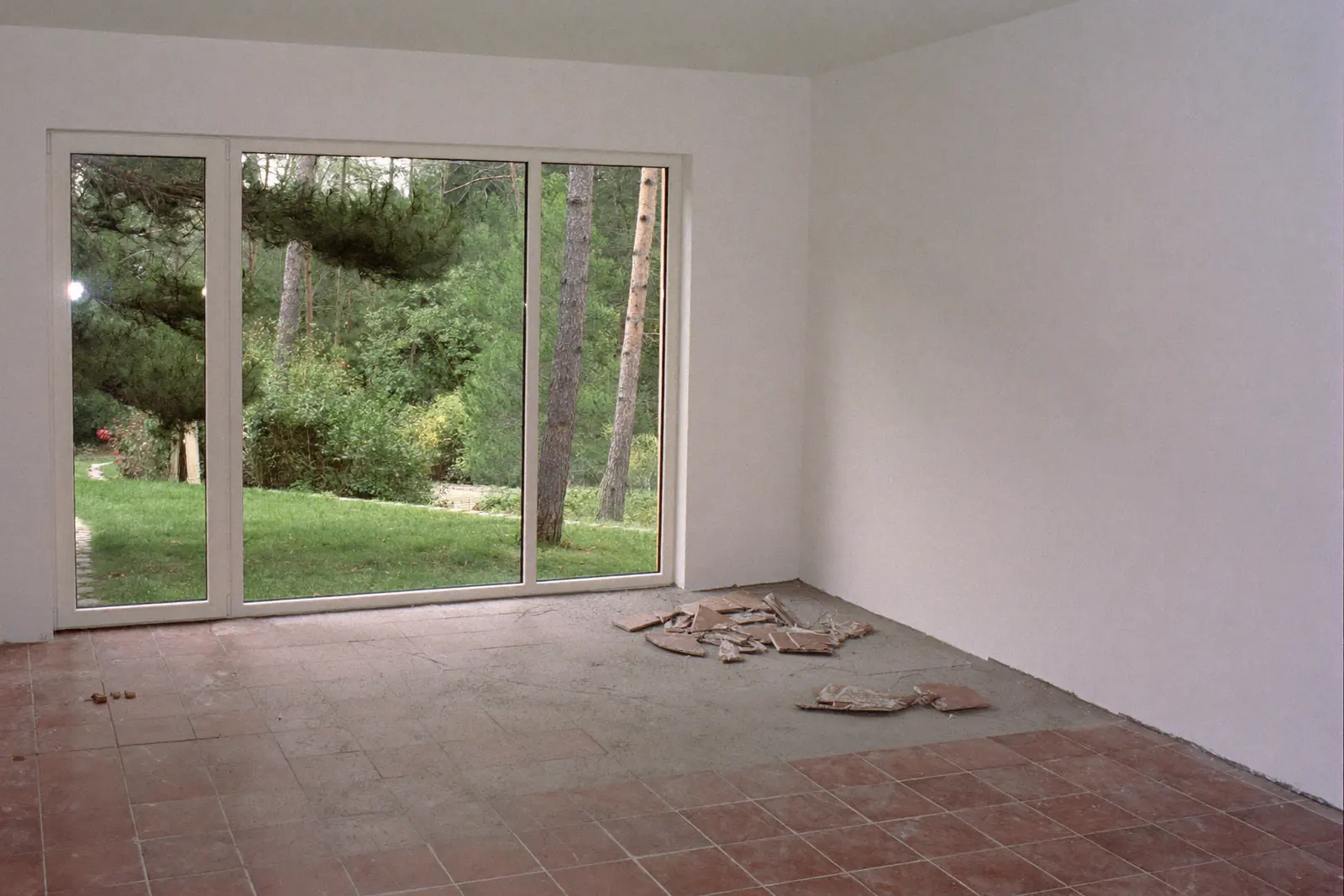 Lege woonkamer met een grote glazen pui naar een tuin met dennen, waar een deel van de oude rode tegels al is verwijderd en de grijze dekvloer zichtbaar is
