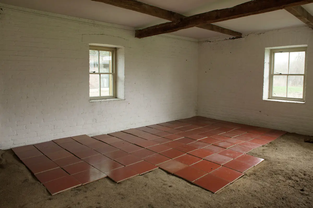Lege kamer in een oude boerderij met witgekalkte muren en houten balken, waar een deel van de rode estrikken al is weggehaald en het zandbed zichtbaar is