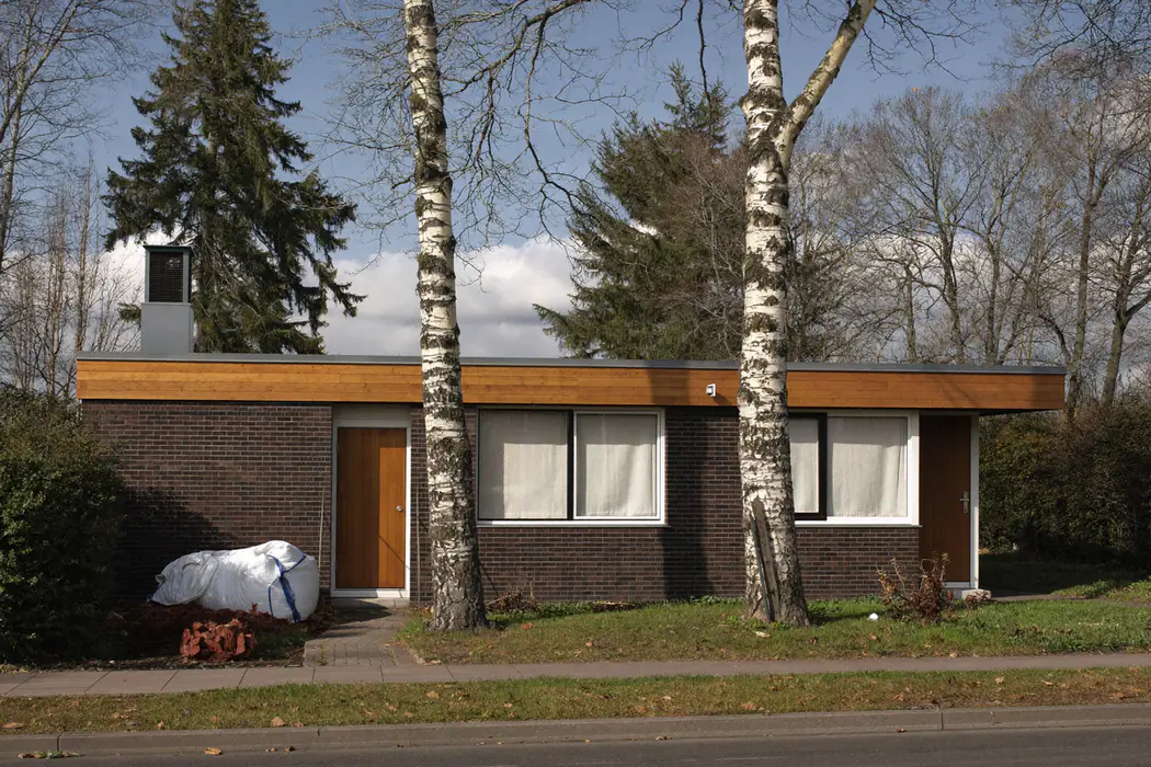 Bungalow met een donkere bakstenen gevel, plat dak en houten boeiboord tussen berken en dennen, met een big bag en oude tegels in de voortuin