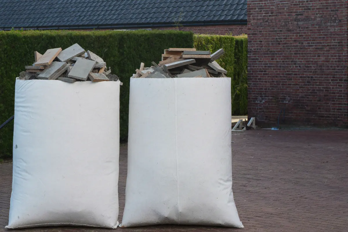Twee witte big bags vol gebroken tegels en puin op een klinkeroprit, voor een haag en een bakstenen gevel
